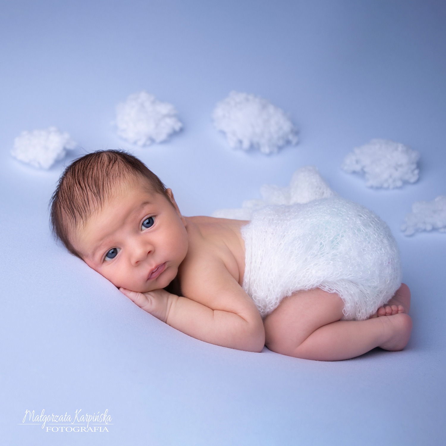 Niemowlę w białym otulaczu leży na boku na jasnoniebieskim tle z dekoracyjnymi białymi obłoczkami, spoglądając w obiektyw.