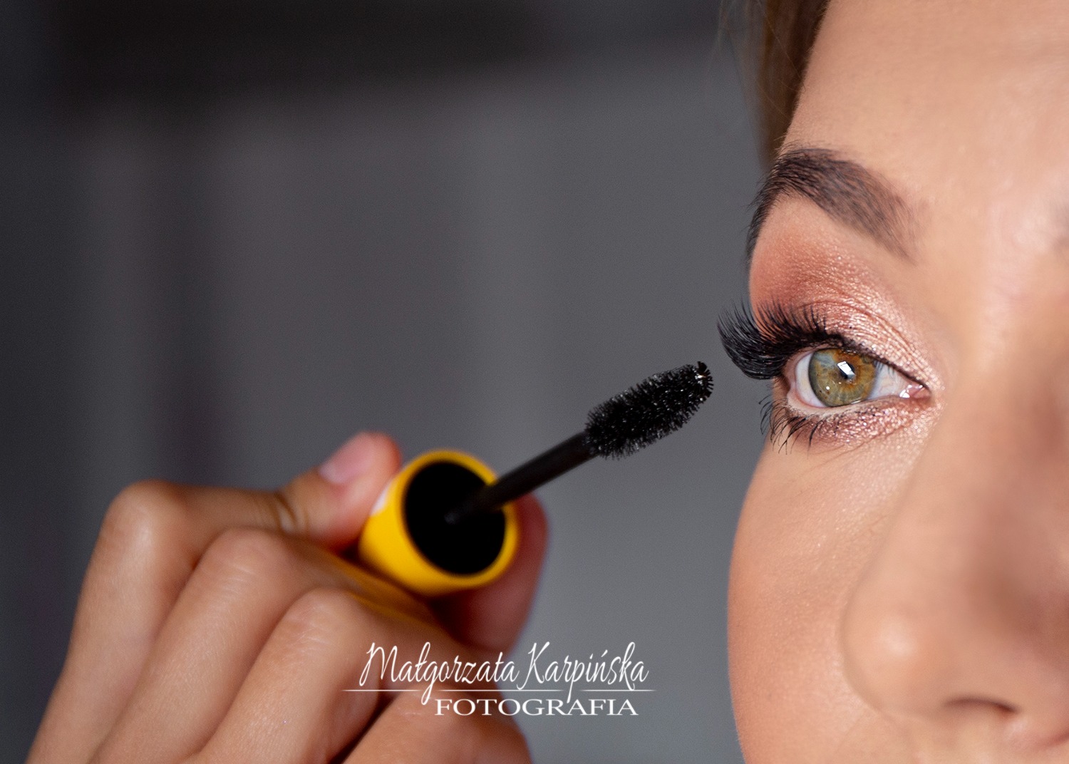 Zbliżenie na oko z długimi rzęsami i błyszczącym cieniem, podczas aplikacji tuszu żółtą szczoteczką.