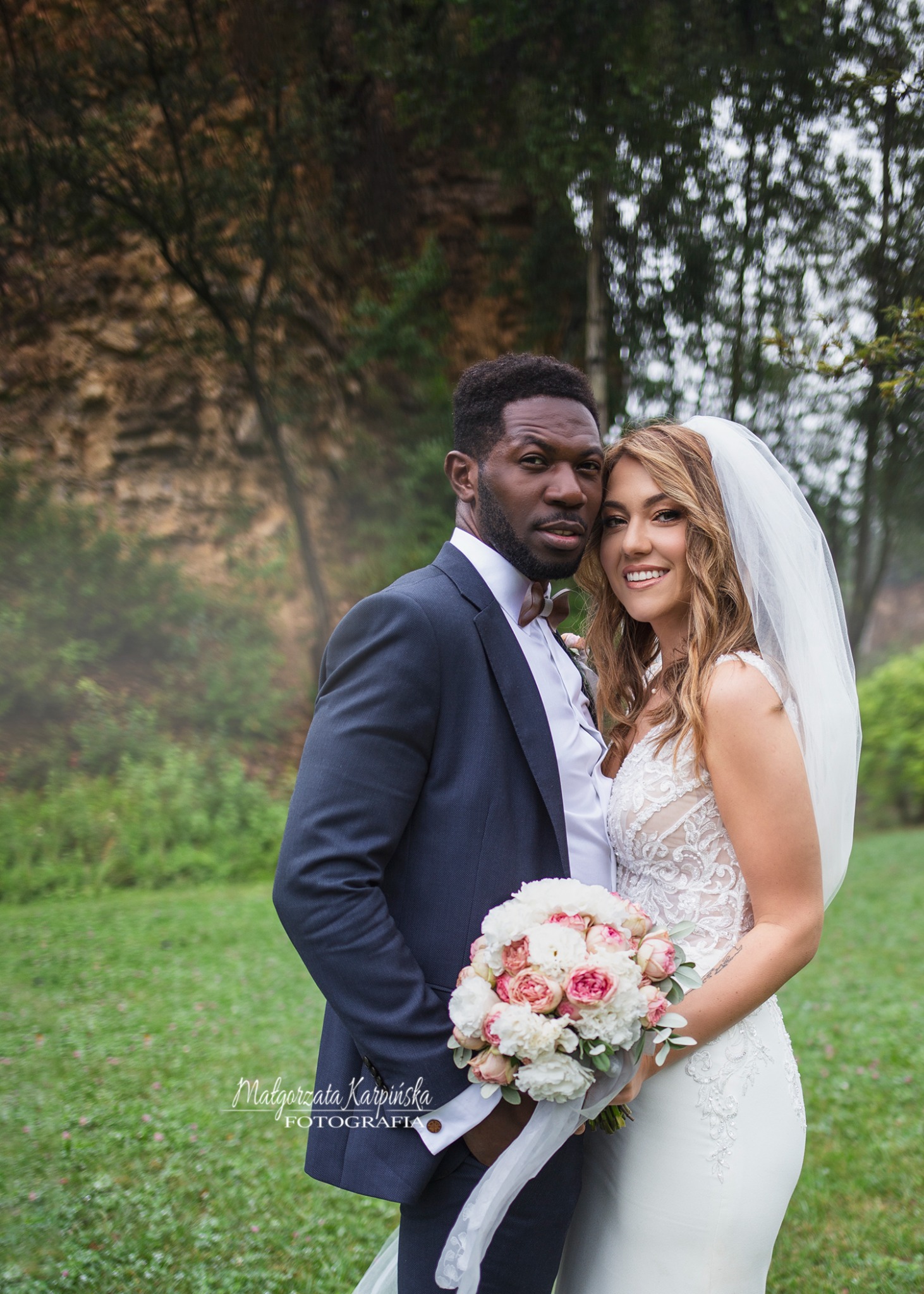 Elegancka para nowożeńców: Afroamerykanin w granatowym garniturze z muchą i panna młoda w koronkowej sukni ślubnej z welonem, trzymająca bukiet różowych i białych kwiatów na tle zielonego trawnika...
