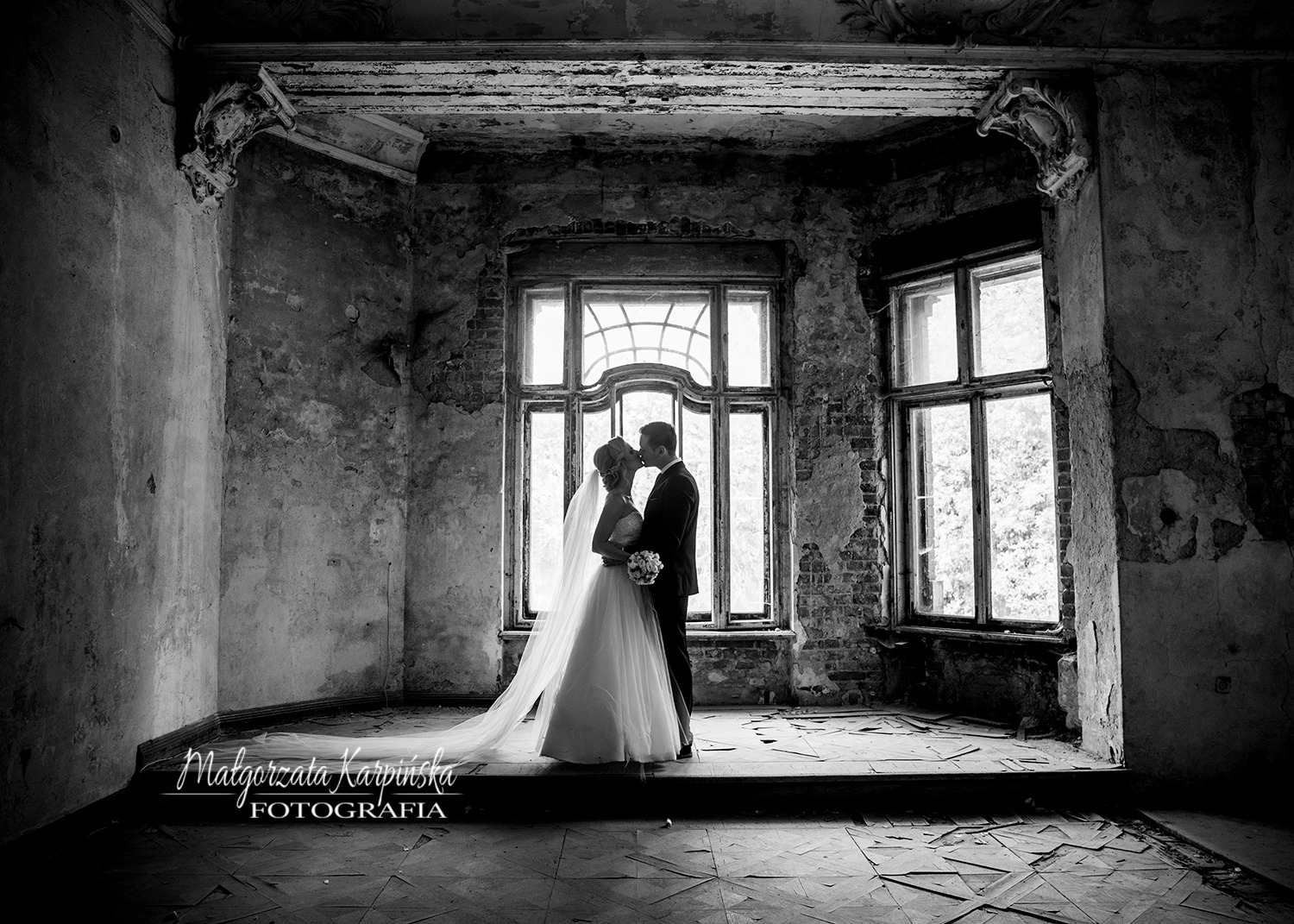 Czarno-białe zdjęcie pary młodej całującej się na tle dużego okna w opuszczonym budynku z odpadającym tynkiem i dekoracyjnymi elementami architektonicznymi.