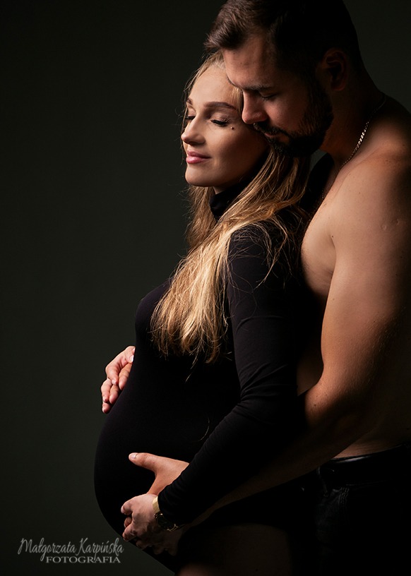 Kobieta w czarnym body z długim rękawem, z zamkniętymi oczami, obejmowana przez mężczyznę bez koszuli, oboje trzymają ręce na jej ciążowym brzuchu, na ciemnym tle.