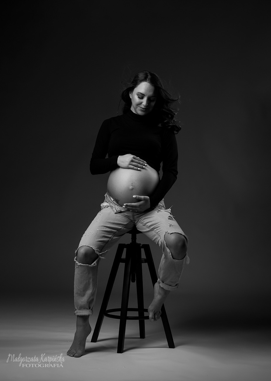 Czarno-białe zdjęcie kobiety w ciąży, siedzącej na stołku, ubranej w czarny golf i podarte jeansy, z rękami na brzuchu, w studiu z ciemnoszarym tłem.