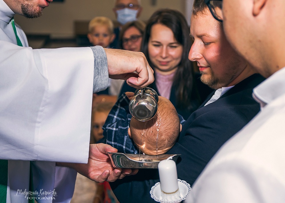 Ksiądz polewa wodą główkę niemowlęcia trzymanego przez ojca podczas ceremonii chrztu, w tle widoczni obserwujący.