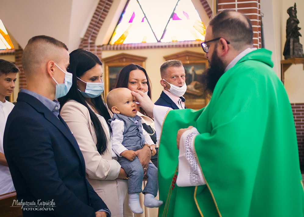 Ksiądz w zielonej szacie dotyka czoła niemowlęcia ubranego w elegancki, szary garnitur podczas uroczystości w kościele, w tle rodzina w maseczkach.