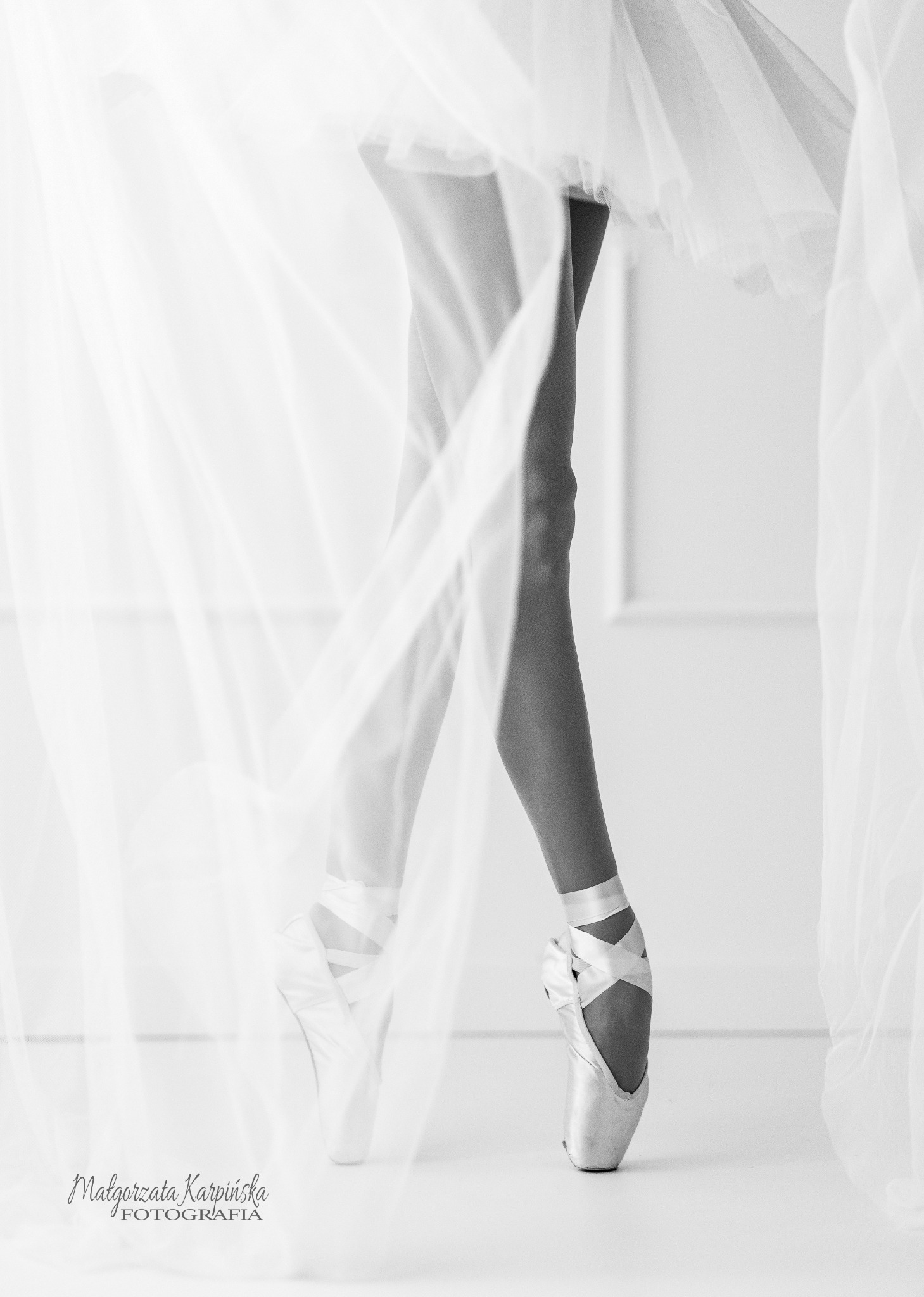 Czarno-białe zdjęcie nóg baletnicy w pointach, otulonych zwiewnym tiulem, na tle minimalistycznego wnętrza.