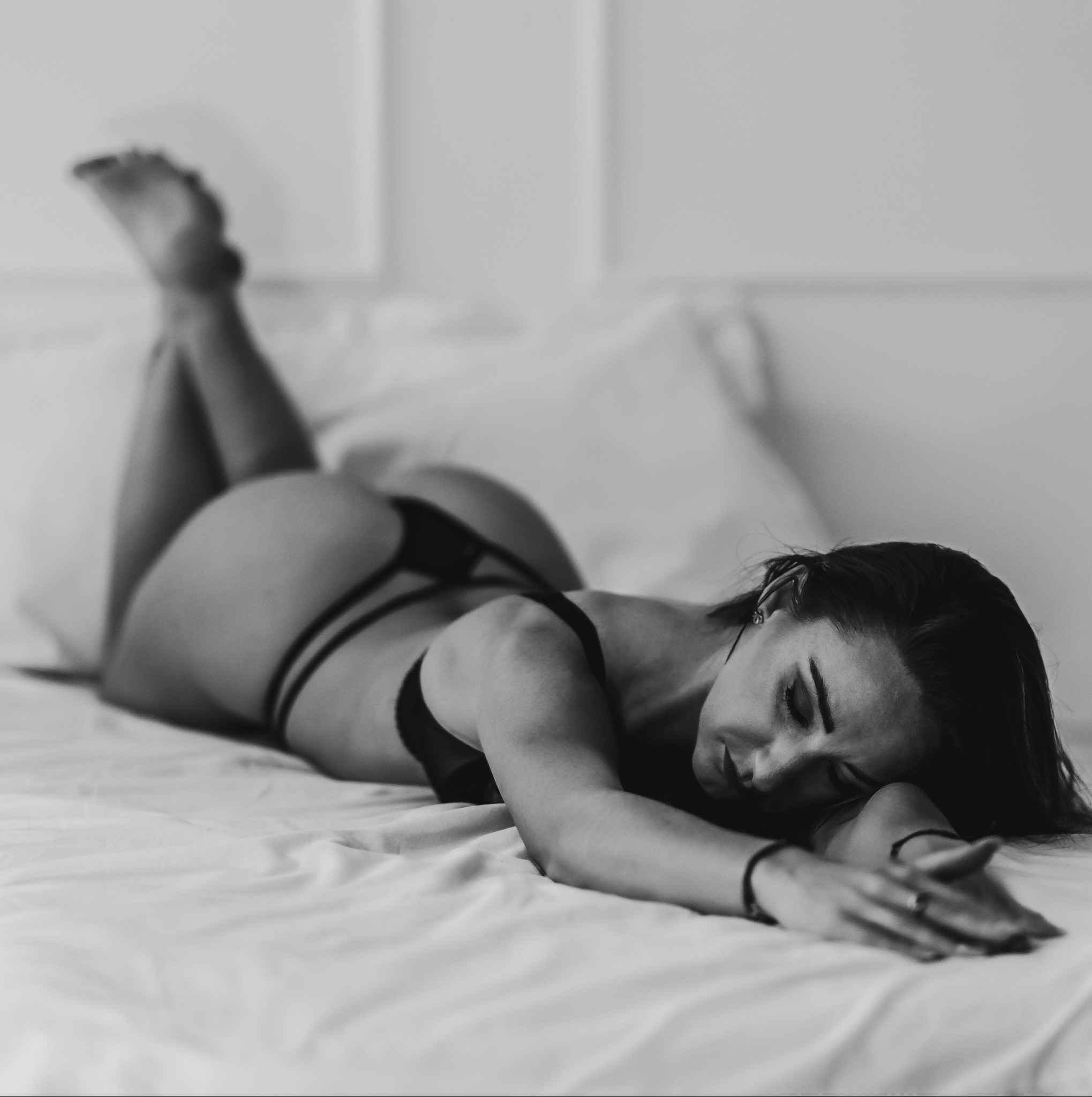 Czarno-białe zdjęcie kobiety leżącej na brzuchu na białej pościeli, z nogami uniesionymi do góry, w delikatnej bieliźnie, z rękami wyciągniętymi przed siebie, zamyślona poza i subtelne oświetlenie.