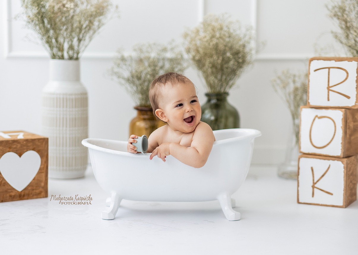 Uśmiechnięty niemowlak w białej wanience, trzymający małą zabawkę, w tle dekoracje z suszonych kwiatów w wazonach i drewniane klocki z literami R, O, K.