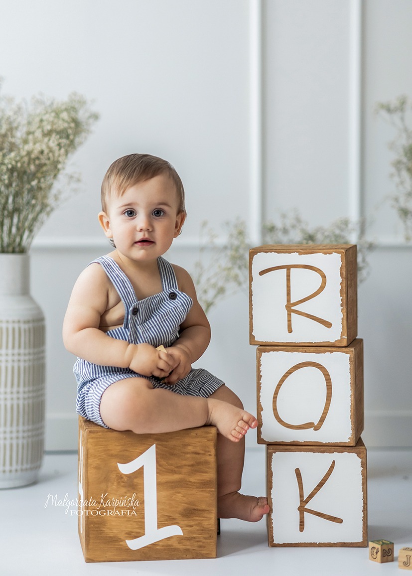 Roczne dziecko w paski siedzi na drewnianym klocku z cyfrą 1, obok wieża z klocków ROK, białe tło, dekoracyjne kwiaty w wazonie.