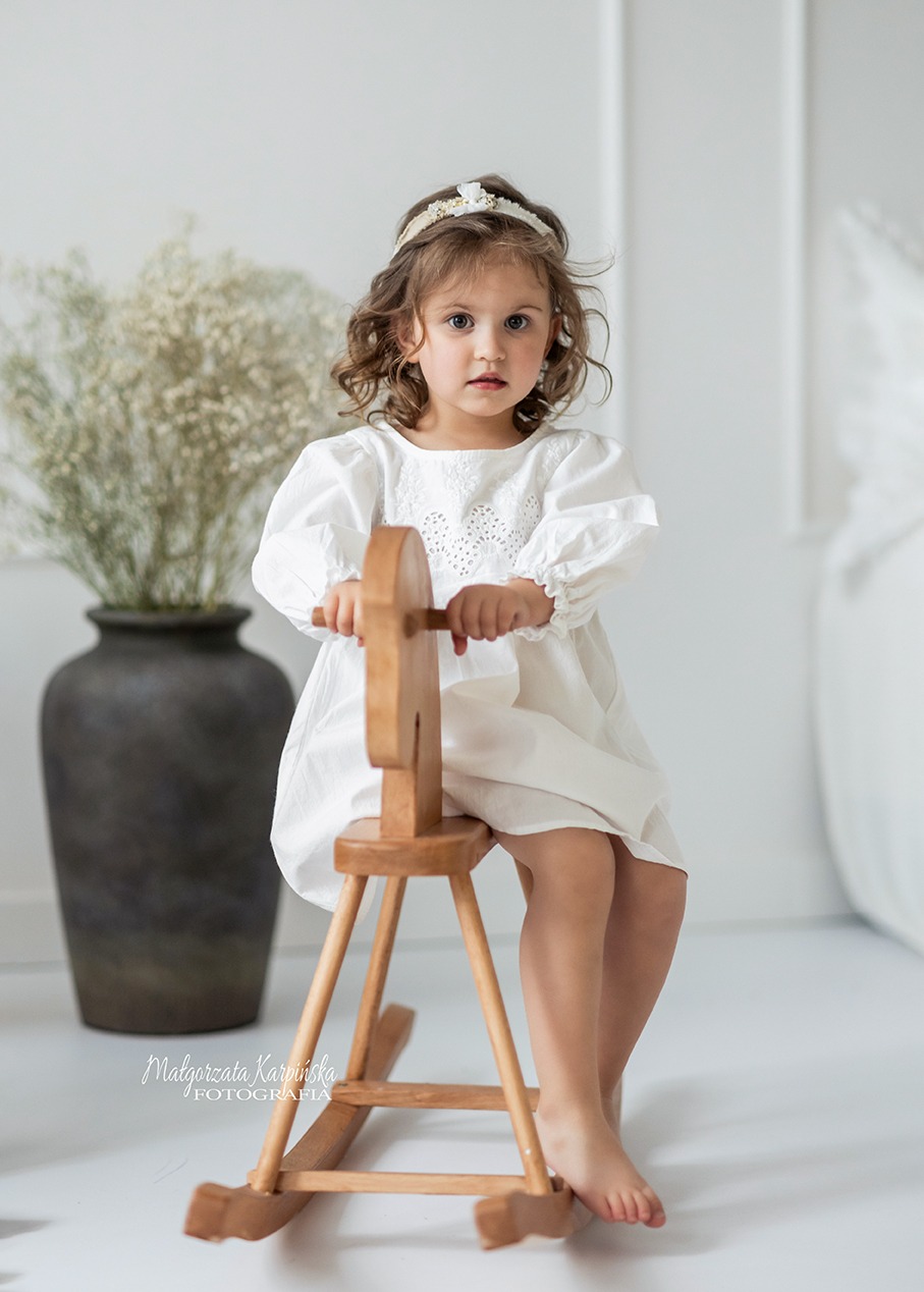 Dziewczynka w białej sukience siedzi na drewnianym koniku na biegunach, w tle wazon z gipsówką i biała ściana.
