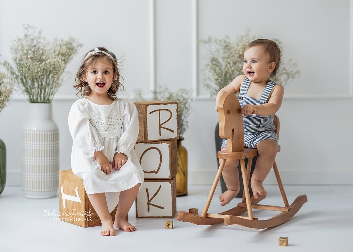 Dwoje uśmiechniętych dzieci: dziewczynka w białej sukience siedzi obok drewnianych klocków z literami R, O, K, a chłopiec w paski siedzi na drewnianym koniku na biegunach w jasnym, minimalistycznym...