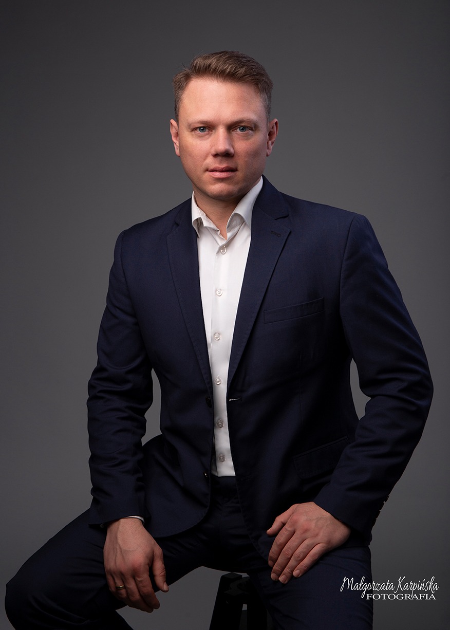 Portret mężczyzny w granatowym garniturze i białej koszuli, siedzącego na stołku na szarym tle, z widocznym pierścionkiem na palcu.