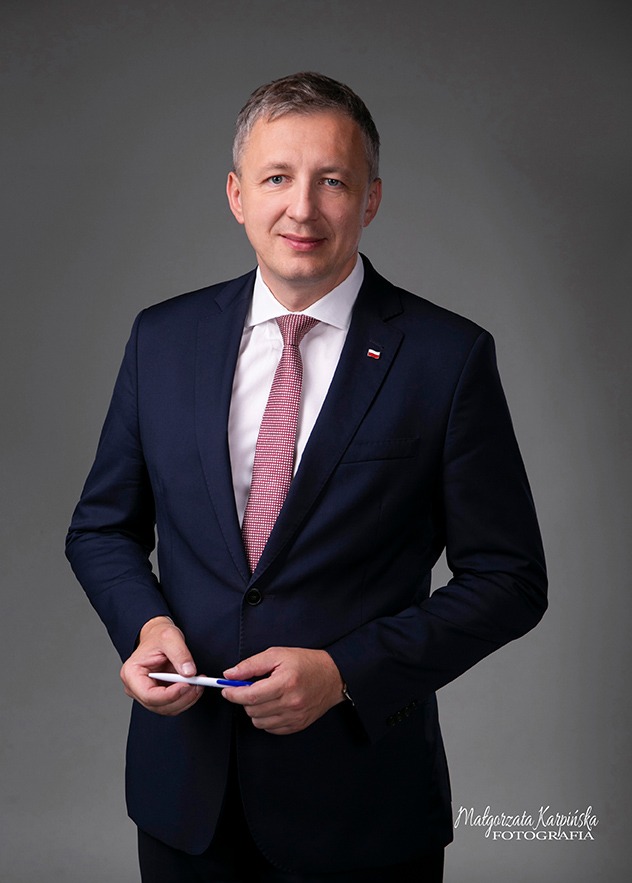 Portret mężczyzny w granatowym garniturze i krawacie z polską flagą w klapie, trzymającego w dłoni długopis na szarym tle.