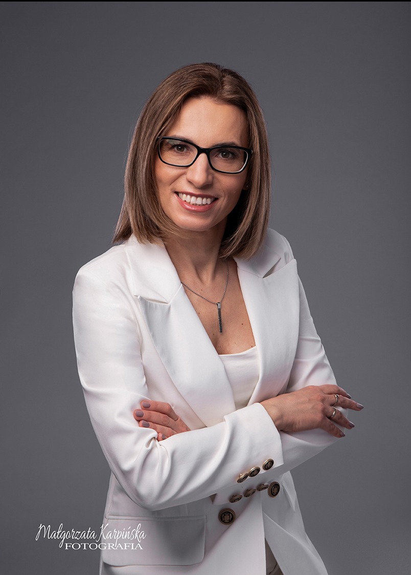 Portret uśmiechniętej kobiety w okularach i białym żakiecie, skrzyżowane ręce, szare tło studyjne, naszyjnik z czarnym wisiorkiem.