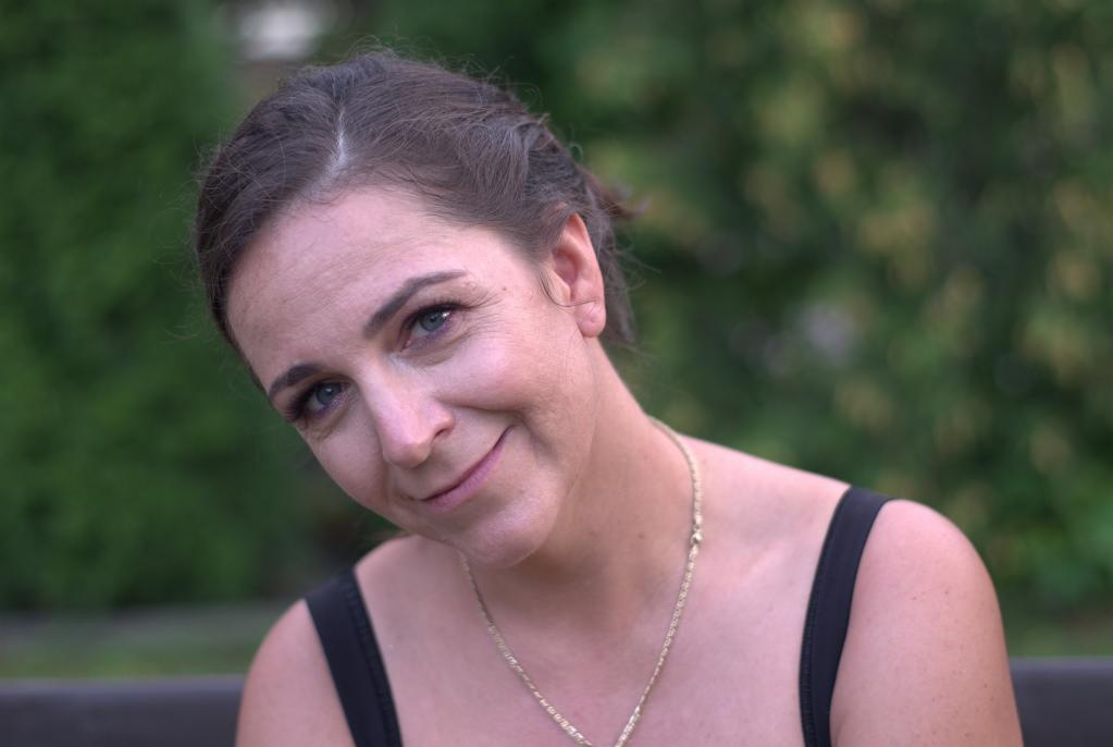 Portret kobiety z uśmiechem, złoty łańcuszek, tło z zielenią.