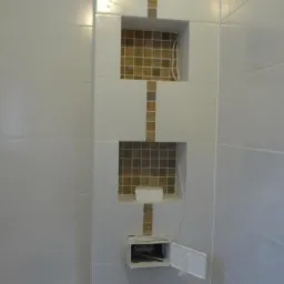 Białe kafelki ścienne z wnękami wykończonymi mozaiką w odcieniach brązu, widoczna otwarta puszka elektryczna z przewodami.