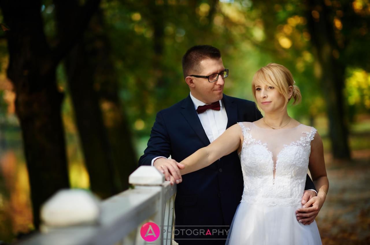 Para młoda w jesiennej scenerii parku: Panna młoda w koronkowej sukni, pan młody w granatowym garniturze i bordowej muszce, opierają się o białą balustradę.