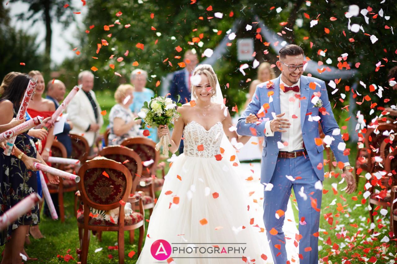 Uśmiechnięta para młoda przechodzi alejką, obsypywana konfetti z czerwonych i białych płatków, goście używają tub z konfetti, on w błękitnym garniturze i bordowej muszce, ona w białej sukni...