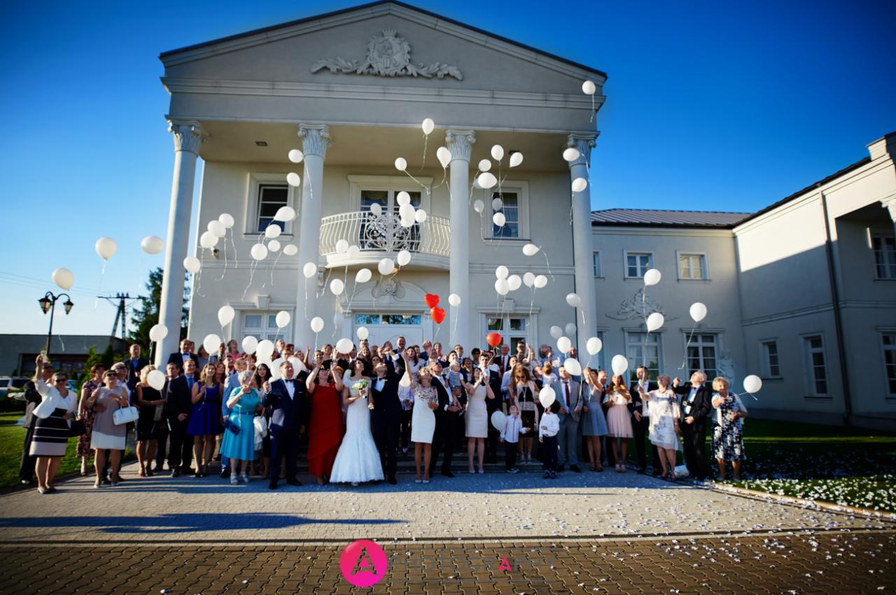 Grupa gości weselnych, w tym para młoda, wypuszcza białe balony przed budynkiem z kolumnami w słoneczny dzień. Dwa czerwone balony wyróżniają się na tle białych.