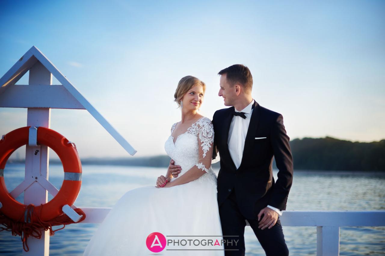 Para młoda w eleganckich strojach pozuje na białym pomoście z kołem ratunkowym, jezioro i las w tle, delikatne oświetlenie.