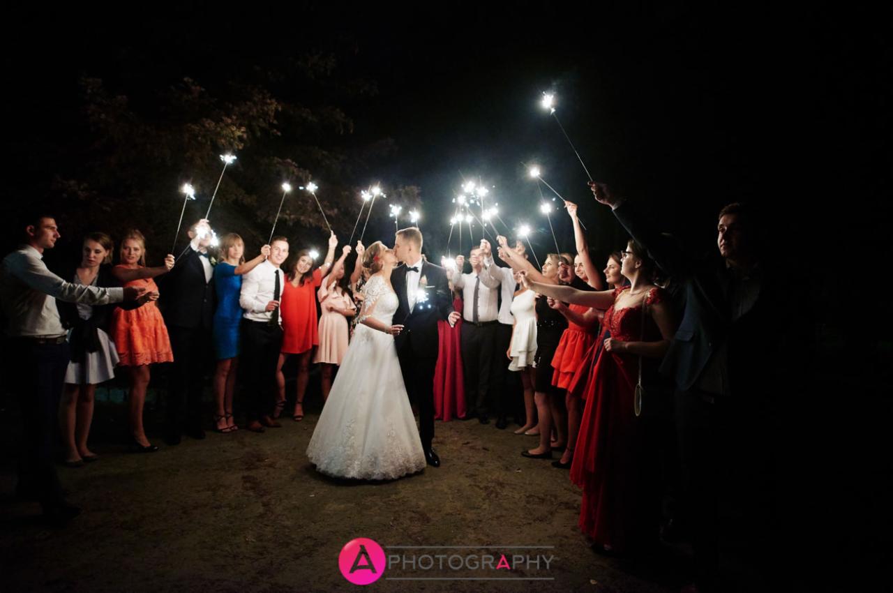 Pocałunek nowożeńców otoczonych gośćmi weselnymi z zimnymi ogniami, nocne ujęcie z ciemnym tłem.