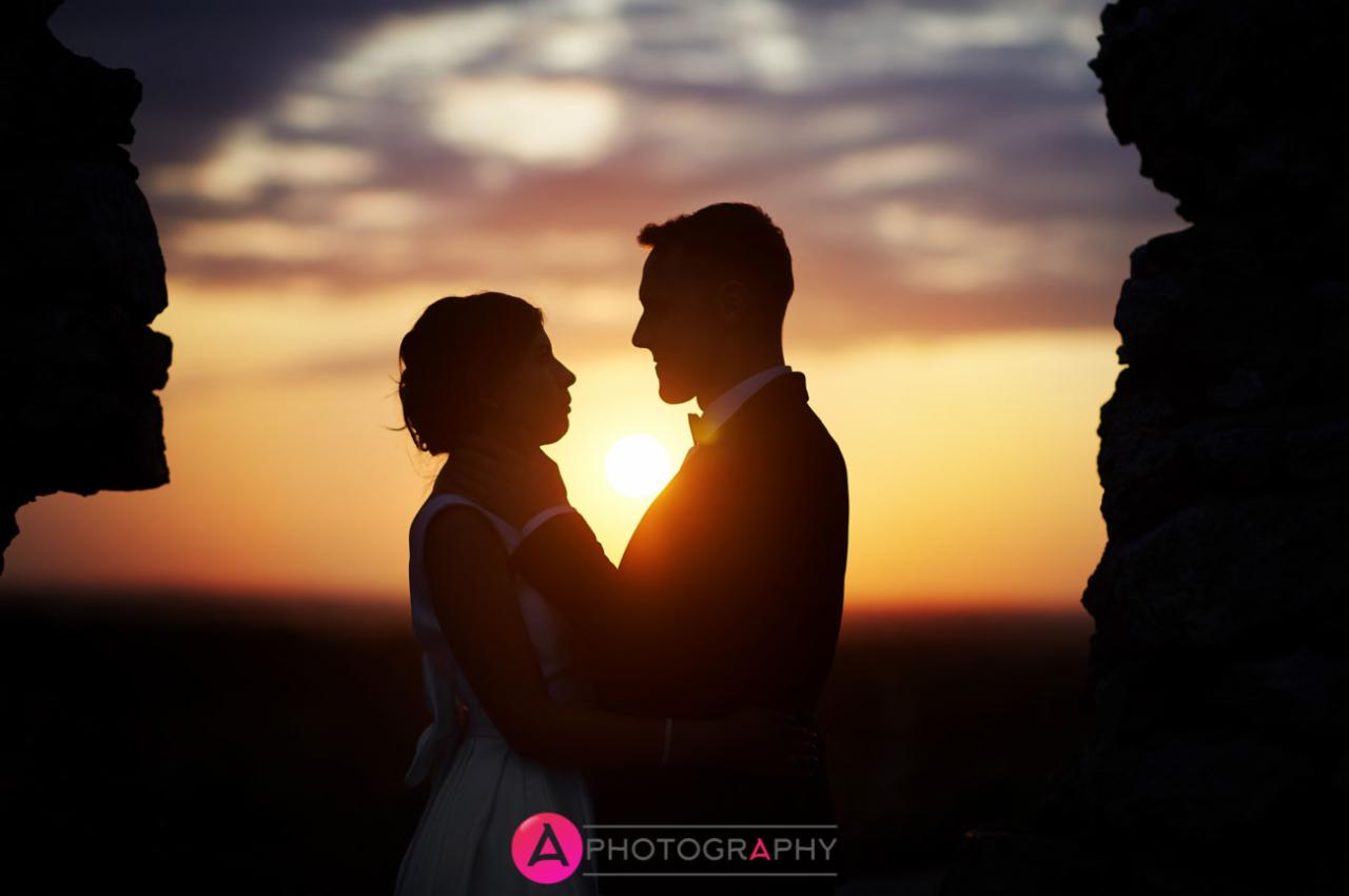 Sylwetki pary młodej na tle zachodzącego słońca, z kamiennymi elementami po bokach kadru.