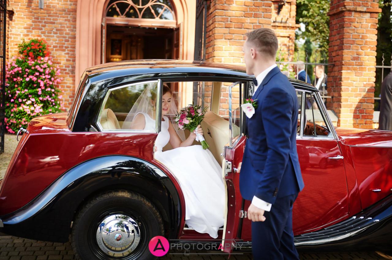 Panna młoda w białej sukni i welonie siedzi w zabytkowym, czerwono-czarnym samochodzie, trzyma bukiet; obok stoi mężczyzna w niebieskim garniturze.