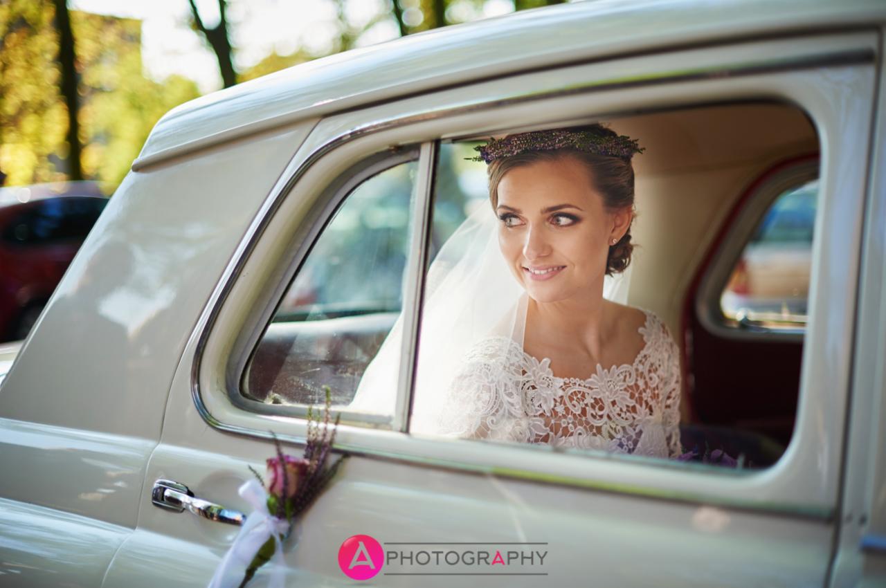 Panna młoda z wiankiem na głowie i welonem spogląda przez okno kremowego retro samochodu, bukiecik z boku.