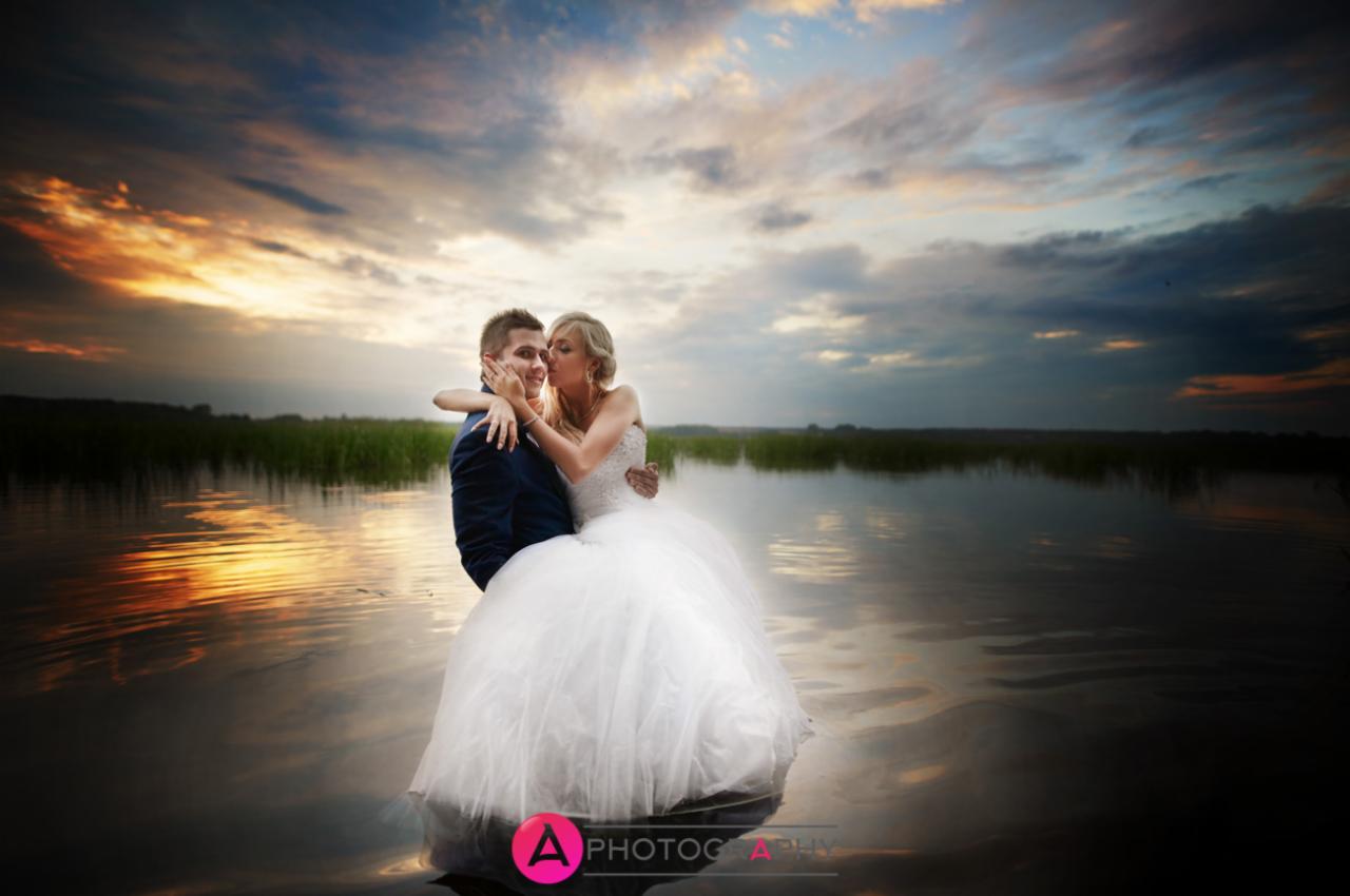 Panna młoda w białej sukni całuje pana młodego w policzek na tle jeziora i zachodzącego słońca.