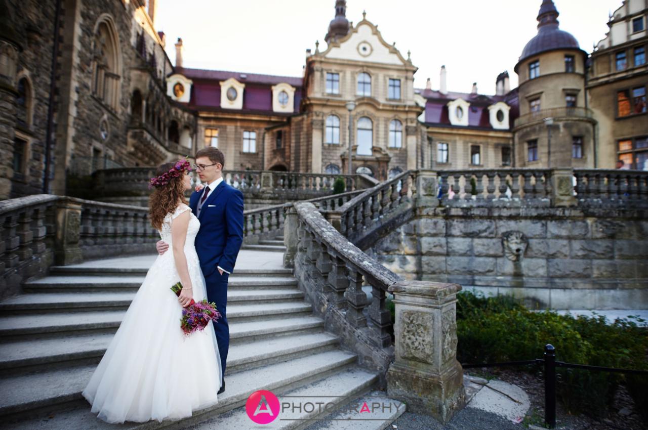 Para młoda na kamiennych schodach zamku, panna młoda z wiankiem z kwiatów i bukietem, pan młody w granatowym garniturze, w tle zamek z wieżyczkami.