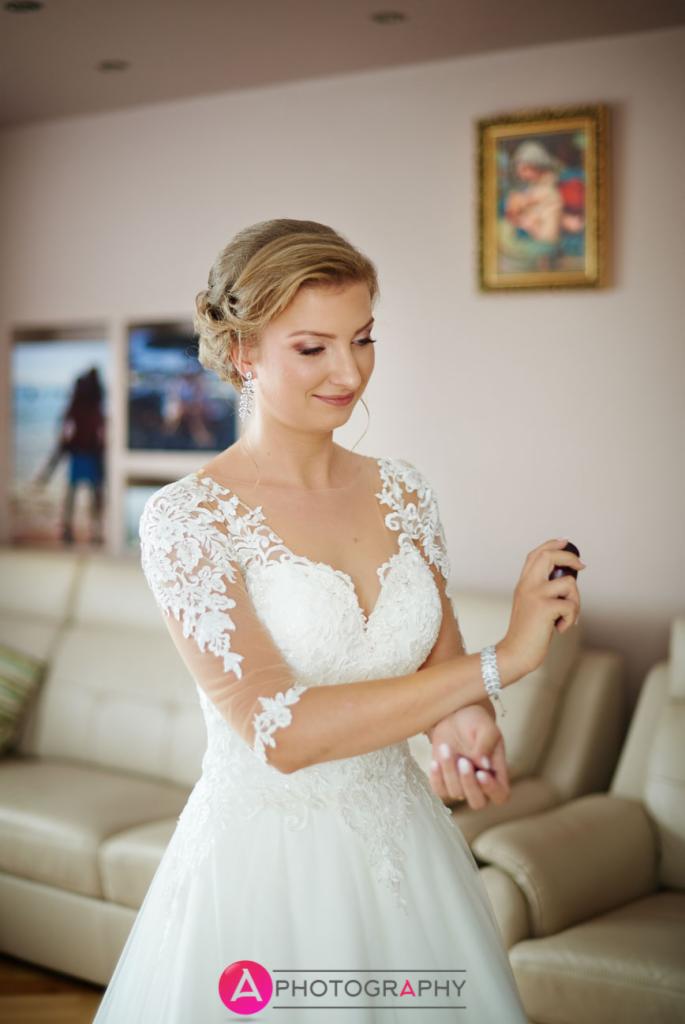 Panna młoda w białej sukni z koronkowymi rękawami i upiętymi włosami spryskuje się perfumami, w tle jasne wnętrze z obrazami.