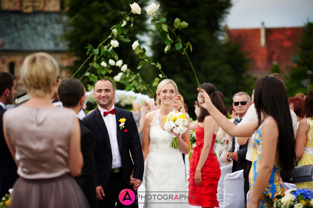 Uśmiechnięta para młoda, trzymająca się za ręce, przechodzi pod łukiem z białych róż, otoczona gośćmi weselnymi na zewnątrz, w tle budynek z czerwonym dachem.