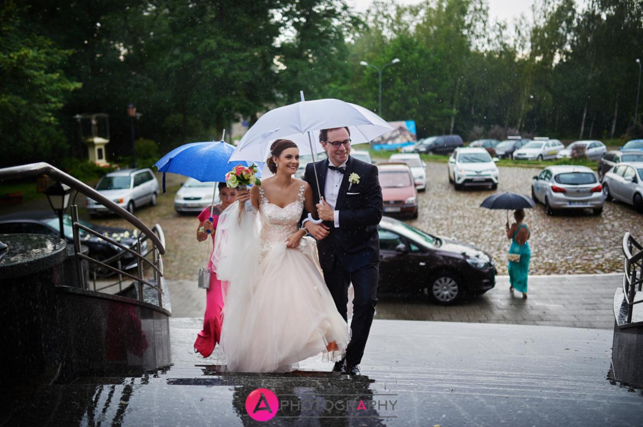 Para młoda w deszczu schodzi po schodach, trzymając biały parasol, w tle parking i drzewa.