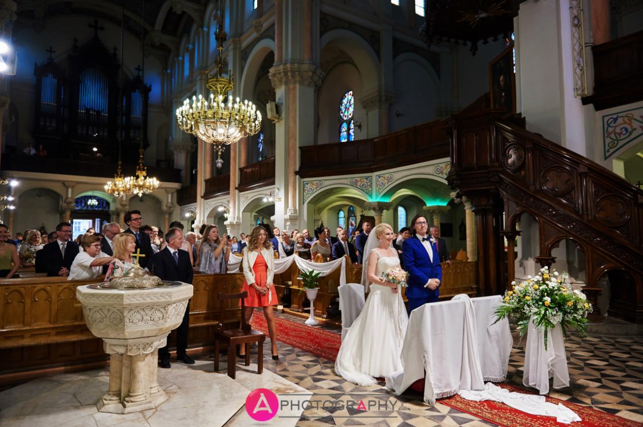 Para młoda stoi przed ołtarzem w kościele, goście obserwują z ławek, w tle organy i witraże.