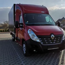 Czerwony Renault Master z plandeką na parkingu z kostki brukowej, widok z przodu, niebo z chmurami w tle.