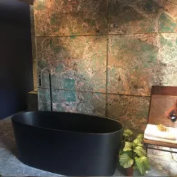 Nowoczesna łazienka z wolnostojącą czarną wanną na tle ściany wyłożonej płytkami imitującymi zielony marmur, obok krzesło z ręcznikami i rośliną doniczkową.