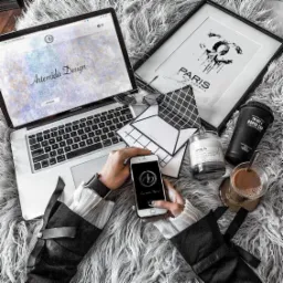 Laptop z otwartą stroną internetową Artemida Design, telefon w dłoniach prezentujący logo firmy, ramka z napisem Paris, świeca, kawa na puszystym tle.