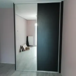 Wnętrze pokoju z zamontowaną szafą przesuwną: jedna część drzwi lustrzana, druga czarna, widoczne kartony i grzejnik w odbiciu lustrzanym.