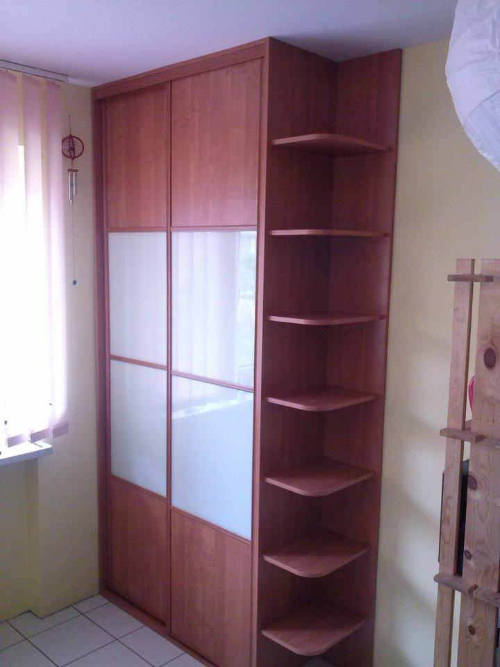 Drewniana szafa przesuwna z białymi, matowymi panelami, połączona z otwartym regałem narożnym z półkami w kolorze drewna, ustawiona w rogu pokoju z jasnymi ścianami i płytkami podłogowymi.