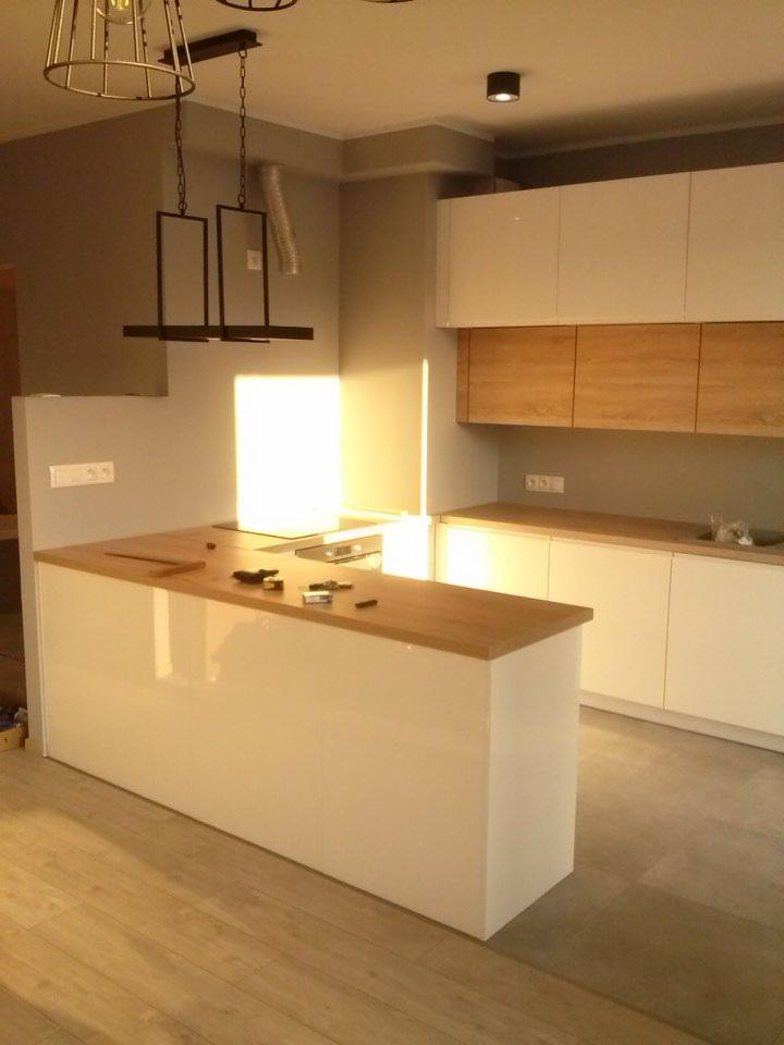 Nowoczesna kuchnia w odcieniach szarości i bieli z drewnianymi akcentami, wyspa z blatem z jasnego drewna, wisząca lampa o geometrycznym kształcie, widoczne narzędzia na blacie.