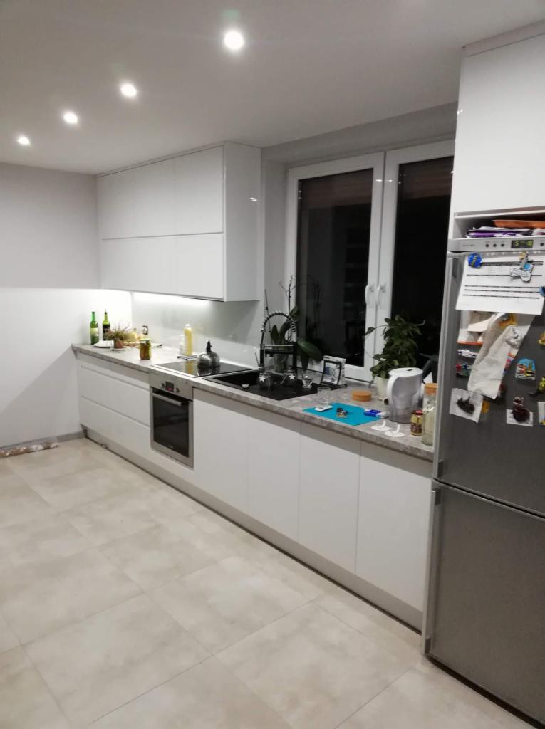 Nowoczesna kuchnia z białymi, gładkimi frontami, szarym blatem i płytkami podłogowymi w odcieniach beżu, widoczne akcesoria kuchenne i roślina w doniczce przy oknie.