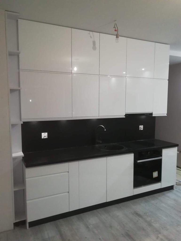 Nowoczesna, biała zabudowa kuchenna z czarnym blatem i czarnym panelem nad blatem, widoczne górne szafki z połyskiem i otwarte półki po lewej stronie, na suficie widoczne wiszące kable od oświetlenia.