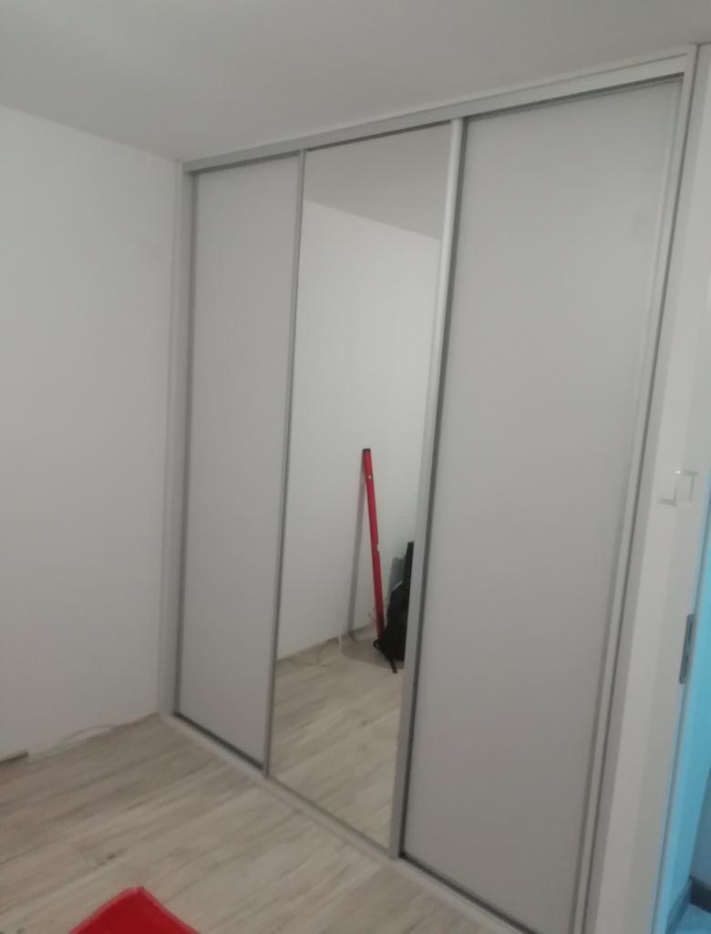 Trzydrzwiowa szafa z przesuwnymi drzwiami, środkowe drzwi lustrzane, widoczna poziomica w odbiciu, na podłodze częściowo widoczny czerwony dywanik.
