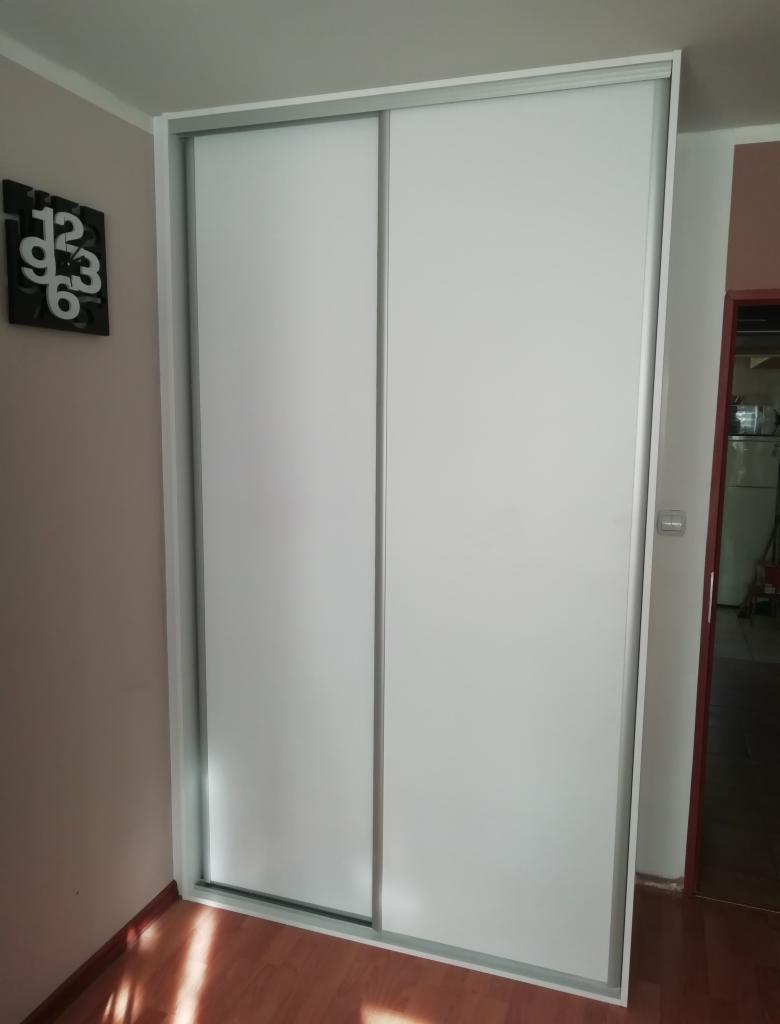 Biała szafa przesuwna z dwoma drzwiami, wbudowana we wnękę, z widocznym zegarem na ścianie po lewej stronie i otwartymi drzwiami do innego pomieszczenia w tle po prawej.