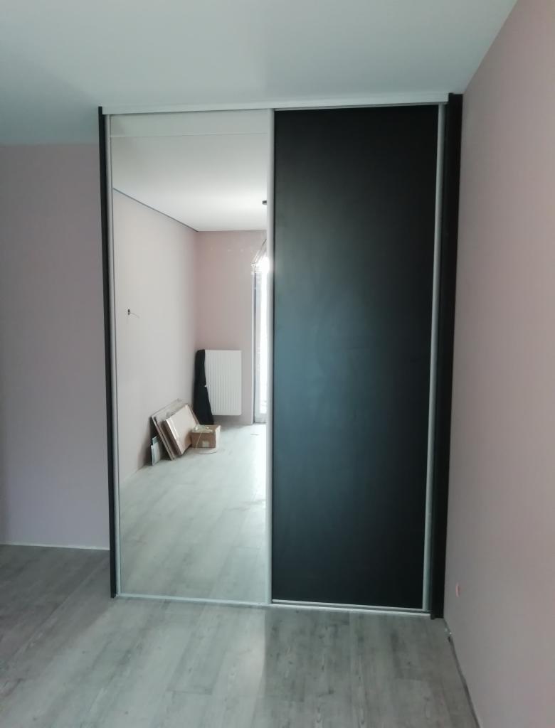 Wnętrze pokoju z zamontowaną szafą przesuwną: jedna część drzwi lustrzana, druga czarna, widoczne kartony i grzejnik w odbiciu lustrzanym.