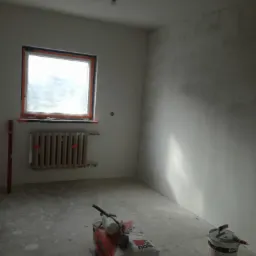 Pokój w trakcie prac przygotowawczych do wykończenia ścian: częściowo wyszpachlowane ściany, zakryte okno folią, grzejnik, paczka gładzi na podłodze, wiadro z zaprawą i narzędzia.