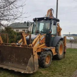 Pomarańczowo-czarna koparko-ładowarka marki CASE stoi na trawie przed domem jednorodzinnym i polami w pochmurny dzień.