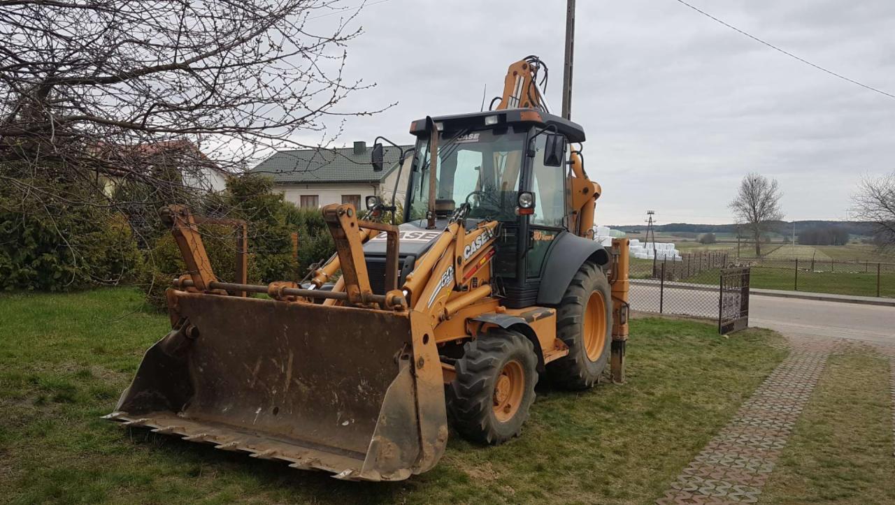 Pomarańczowo-czarna koparko-ładowarka marki CASE stoi na trawie przed domem jednorodzinnym i polami w pochmurny dzień.