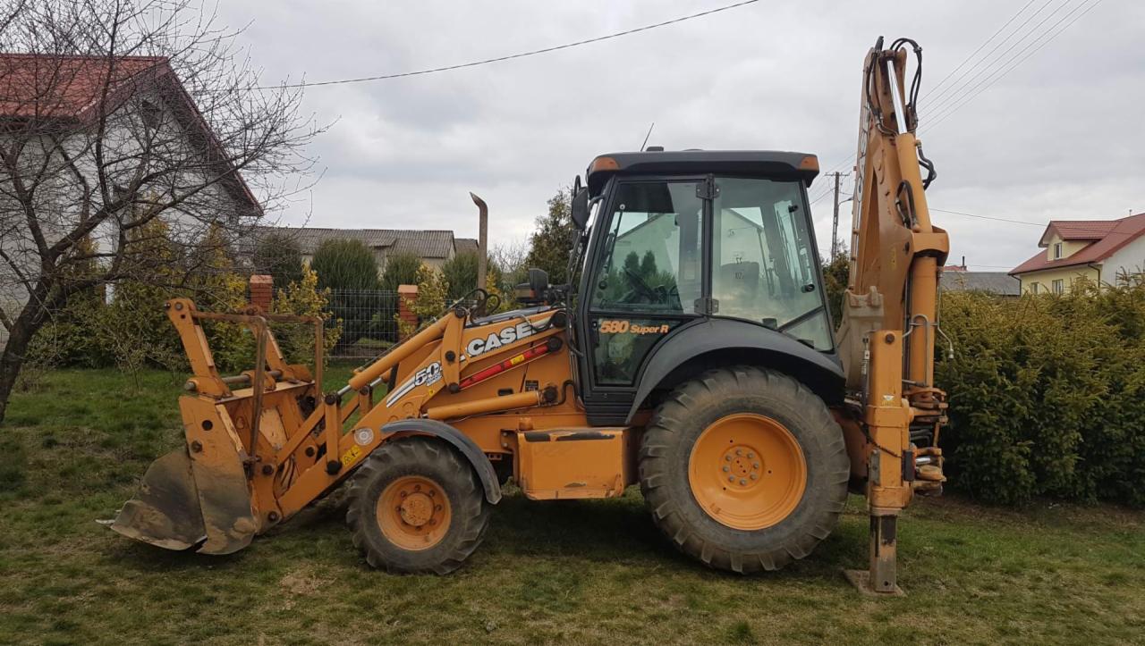 Żółta koparko-ładowarka Case 580 Super R stoi na trawie przed domem jednorodzinnym w pochmurny dzień.