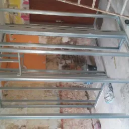 Metalowa konstrukcja szkieletowa na ścianie w surowym stanie, przygotowana do montażu płyt, z widocznymi fragmentami starej cegły i narzędziem szlifierskim leżącym na podłodze.