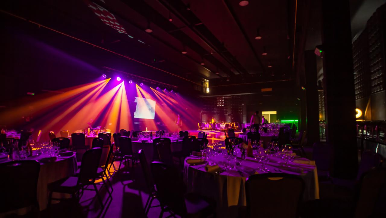 Sala bankietowa przygotowana na uroczystość, oświetlona fioletowymi i pomarańczowymi reflektorami, z nakrytymi stołami i ustawionymi krzesłami, gotowa na przyjęcie gości.